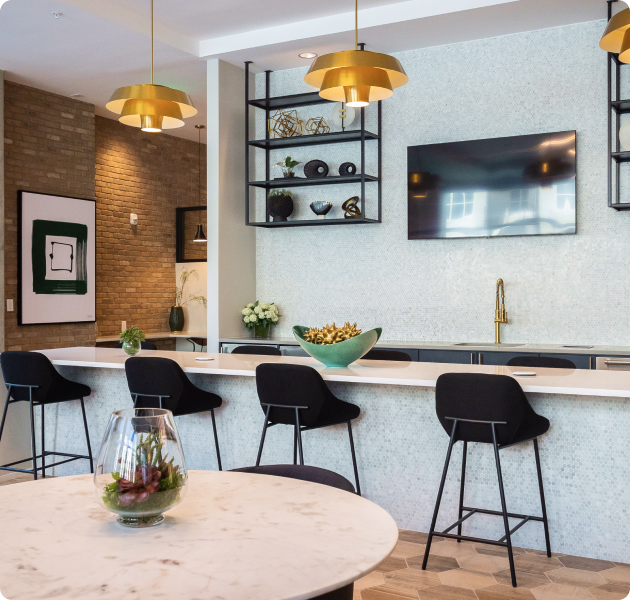 A community center features a bar counter with sleek black barstools, three elegant gold pendant lights and a wall-mounted TV positioned above a luxurious gold faucet at MAA Optimist Park luxury apartments in Charlotte, NC