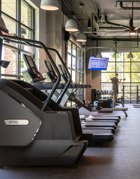 A modern fitness center equipped with stair master and treadmills next to large picture windows, with a wall-mounted TV and overhead lighting at MAA Optimist Park luxury apartments in Charlotte, NC