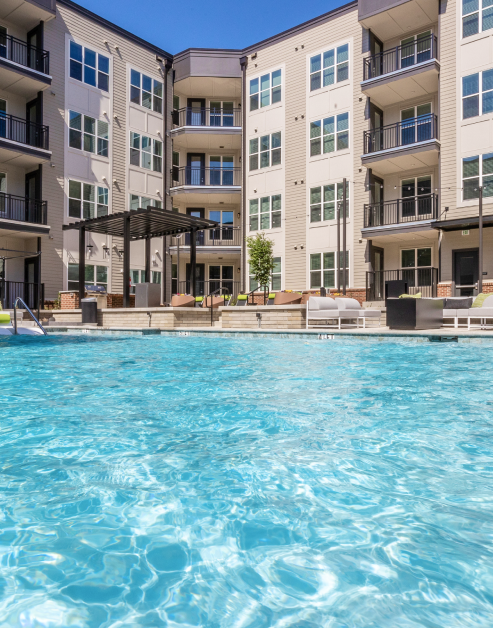 Luxurious swimming pool surrounded by apartment complex with lounge chairs and umbrellas, next to shaded pergola seating area at MAA Optimist Park luxury apartments in Charlotte, NC