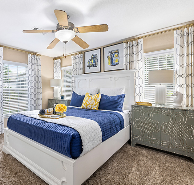 An apartment model bedroom with ceiling fan and wall paintings, queen bed with plush blue comforter with windows covered with blinds and curtains at MAA Prosperity Creek luxury apartments in Charlotte, NC