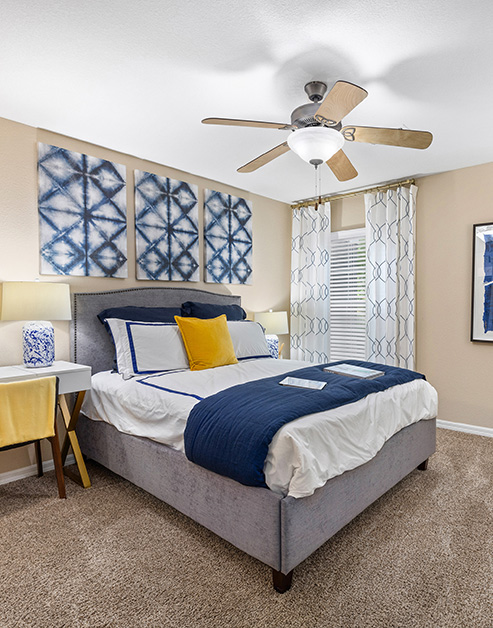 Model bedroom with ceiling fan, wall paintings above bed next to a window with blinds and curtains at MAA Prosperity Creek luxury apartments in Charlotte, NC
