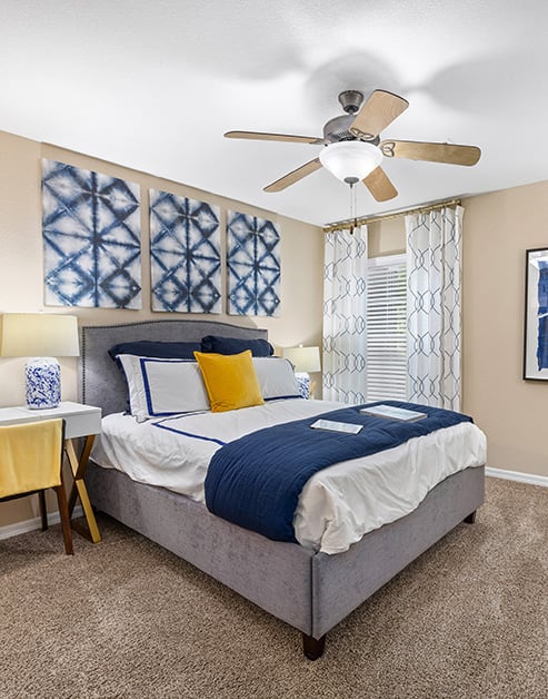 Model bedroom with ceiling fan, wall paintings above bed next to a window with blinds and curtains at MAA Prosperity Creek luxury apartments in Charlotte, NC
