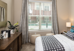 Side view of bedroom with wooden dresser next to the queen bed and windows with blinds in natural light at MAA Reserve luxury apartments in Charlotte, NC
