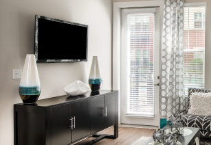 Model living room wall mounted TV above dark cabinet next to the French door, windows with blinds in natural light at MAA Reserve luxury apartments in Charlotte, NC