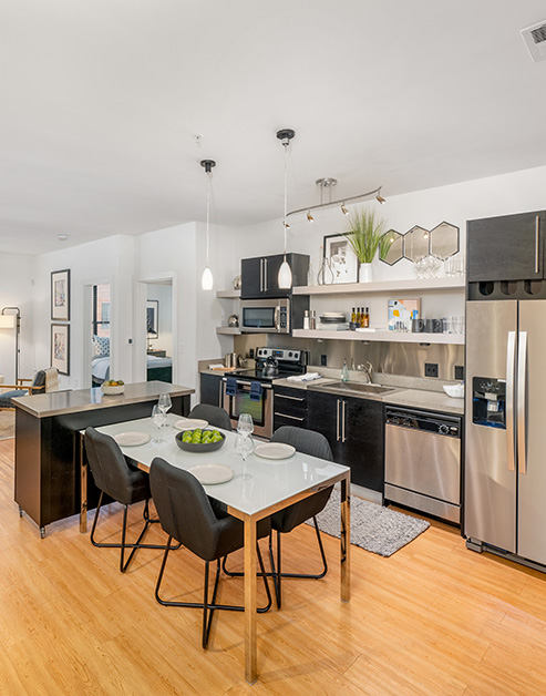 Open-style kitchen with sleek hanging light fixtures, stainless steel appliances, an island countertop and a small dining table on wood floors at MAA South Line luxury apartments in Charlotte, NC