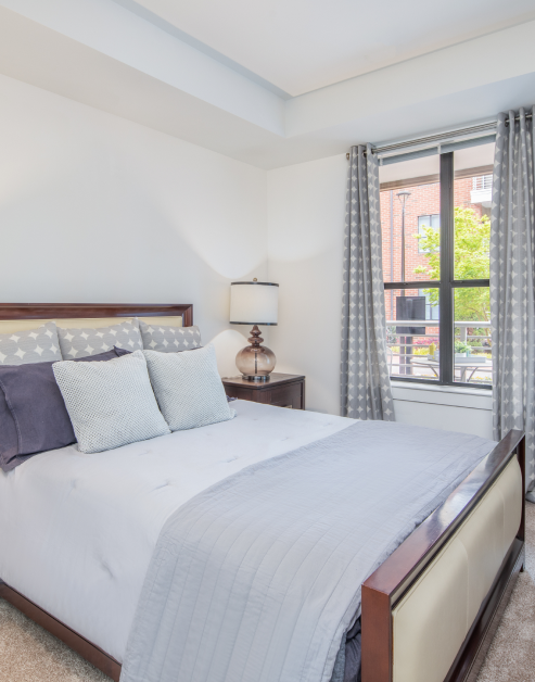 Bedroom bed with light-toned accent pillows, a bedside nightstand with lamp next to window with curtains at MAA South Line luxury apartments in Charlotte, NC