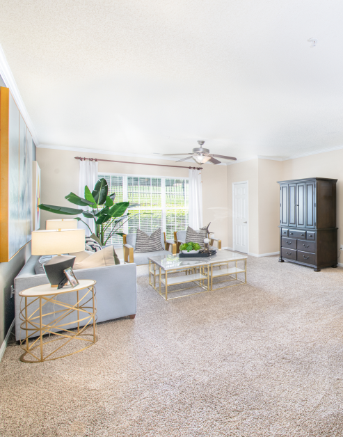 Spacious model living room with a ceiling fan, modern-style couch and rectangle coffee table next to a large picture window with blinds and curtains at MAA University Lake luxury apartments in Charlotte, NC