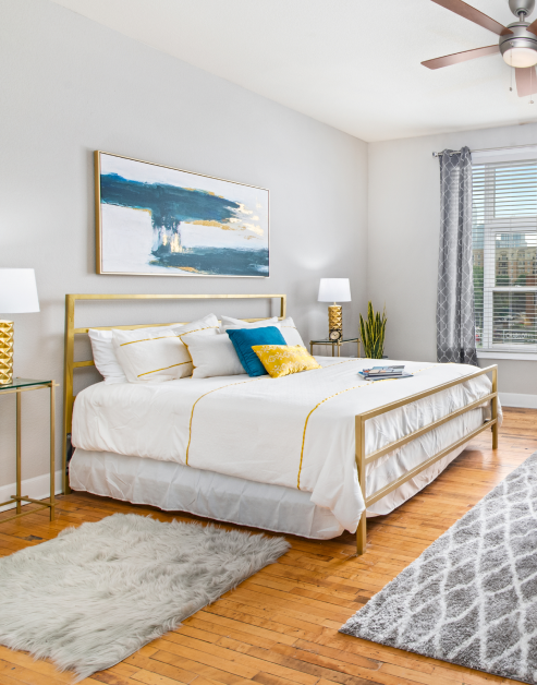 Bedroom with white bed and gold bed frame, a ceiling fan and wall paintings, with area rugs on wood floors at MAA Uptown luxury apartments in Charlotte, NC