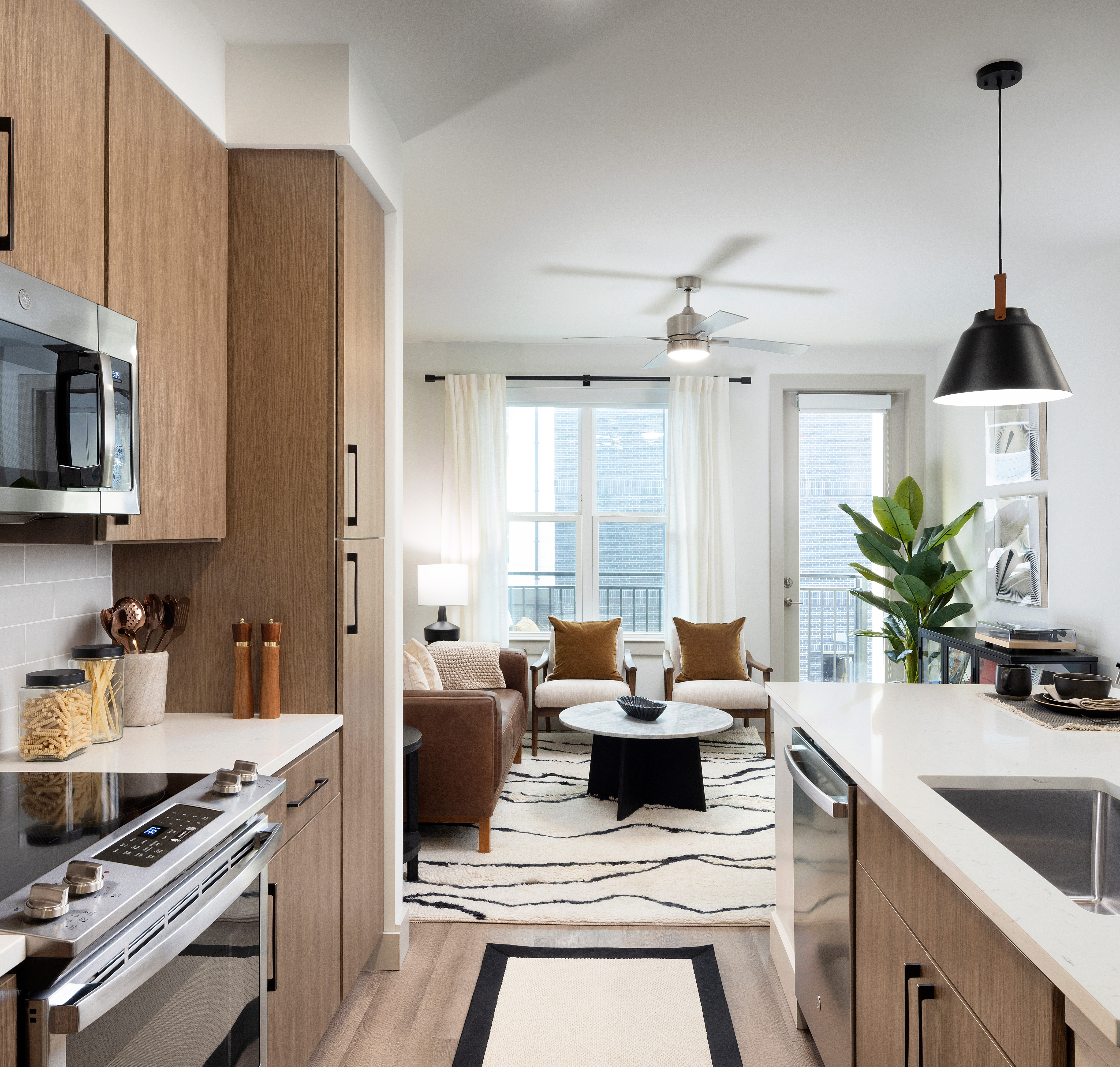 Apartment open kitchen with stainless-steel appliances, light brown wood cabinets and wood floors next to the living area with brown leather couch, armchairs and coffee table on a white area rug at MAA Nixie luxury apartments in Durham, NC