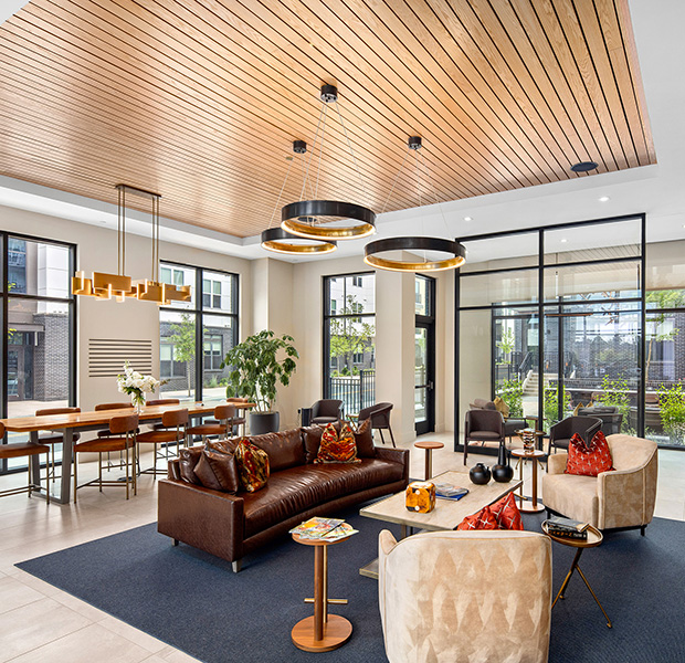 Upscale community clubhouse with a wood beam ceiling, stylish hanging light fixtures above lounge sitting area on an area rug, next to large conference table with chairs surrounded by floor length picture windows at MAA Nixie luxury apartments in Durham, NC