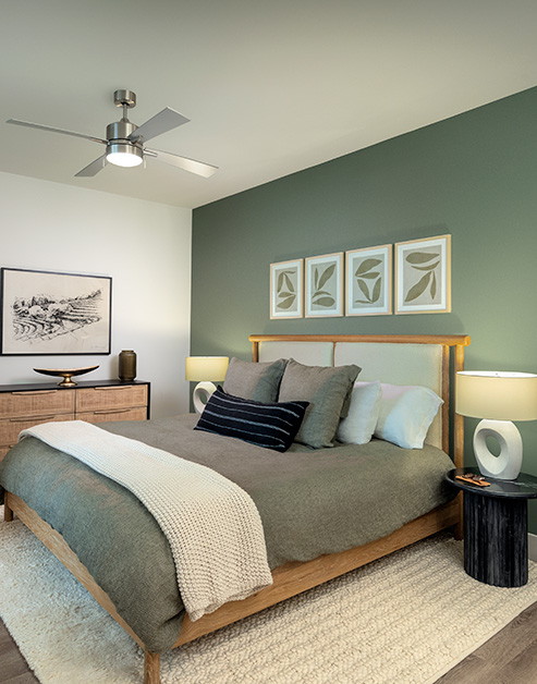 Bedroom with a ceiling fan, green accent wall with framed art above wooden bed frame with an area rug on wood floors at MAA Nixie luxury apartments in Durham, NC