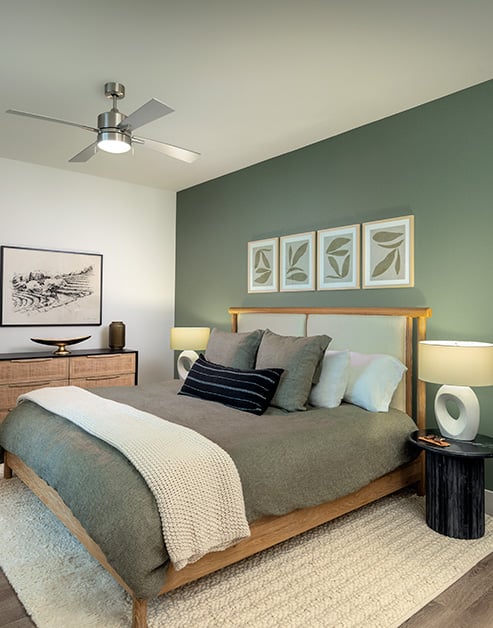 Bedroom with a ceiling fan, green accent wall with framed art above wooden bed frame with an area rug on wood floors at MAA Nixie luxury apartments in Durham, NC