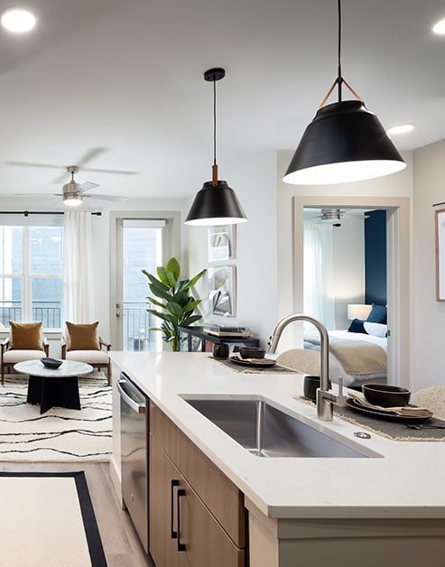 Modern open kitchen with black hanging light fixtures above island with white granite countertops and sink next to living area at MAA Nixie luxury apartments in Durham, NC