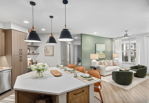 Spacious living room area with a ceiling fan, a large wall painting on sage green accent wall with a white sofa and dark green armchairs next to a white round coffee table on wood floors next to the open kitchen at MAA Nixie luxury apartments in Durham, NC