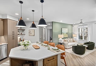 Spacious living room area with a ceiling fan, a large wall painting on sage green accent wall with a white sofa and dark green armchairs next to a white round coffee table on wood floors next to the open kitchen at MAA Nixie luxury apartments in Durham, NC