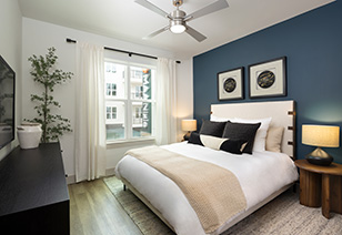 Modern bedroom with a ceiling fan, wall art on blue accent wall above white bedding with accent pillow next to a window with open white curtains at MAA Nixie luxury apartments in Durham, NC
