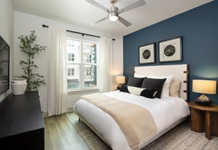 Modern bedroom with a ceiling fan, wall art on blue accent wall above white bedding with accent pillow next to a window with open white curtains at MAA Nixie luxury apartments in Durham, NC