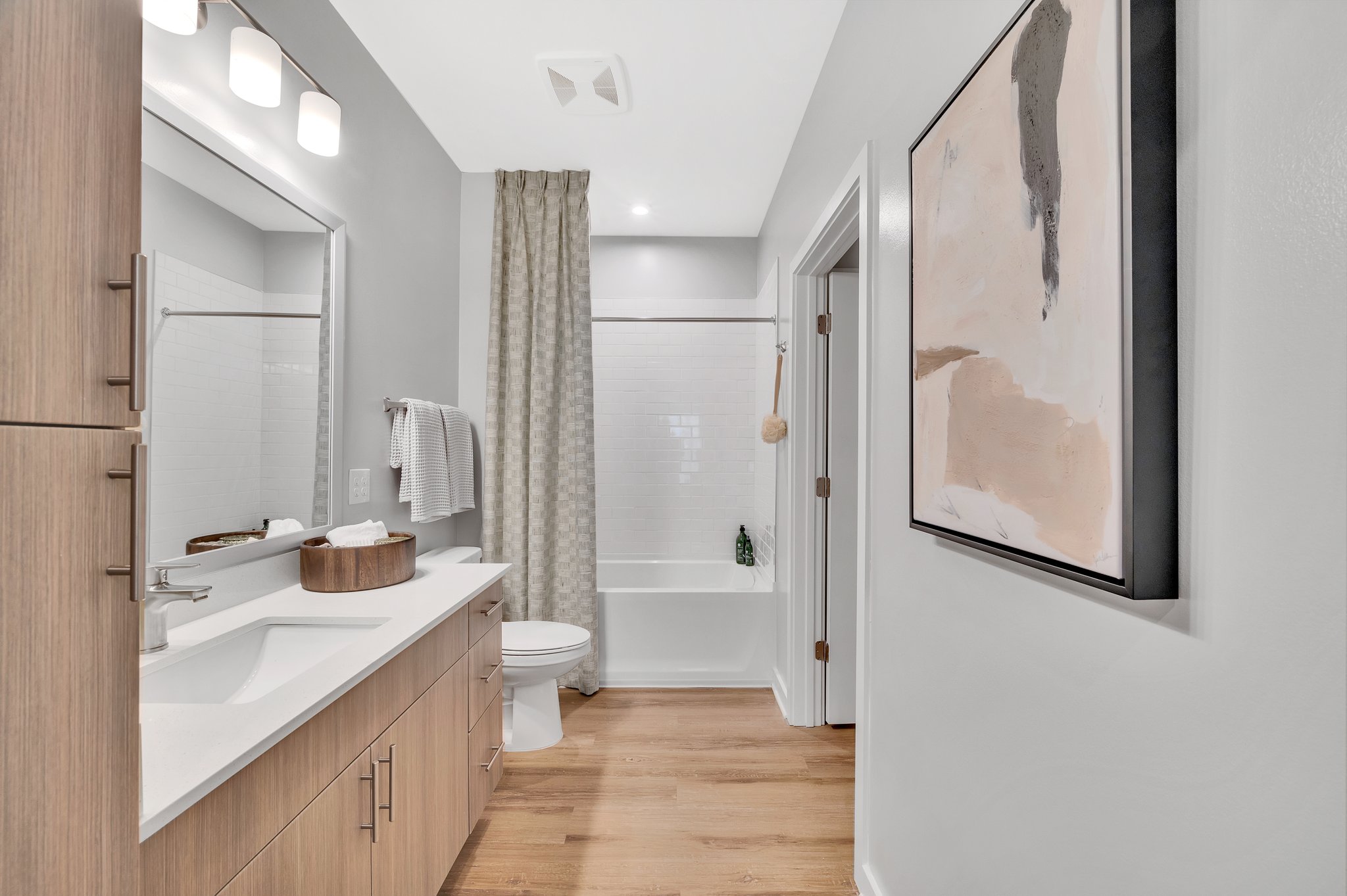 Bathroom with sleek light fixtures above a framed vanity mirror and sink with light brown cabinets and grey shower curtains at MAA Vale luxury apartments in Raleigh, NC