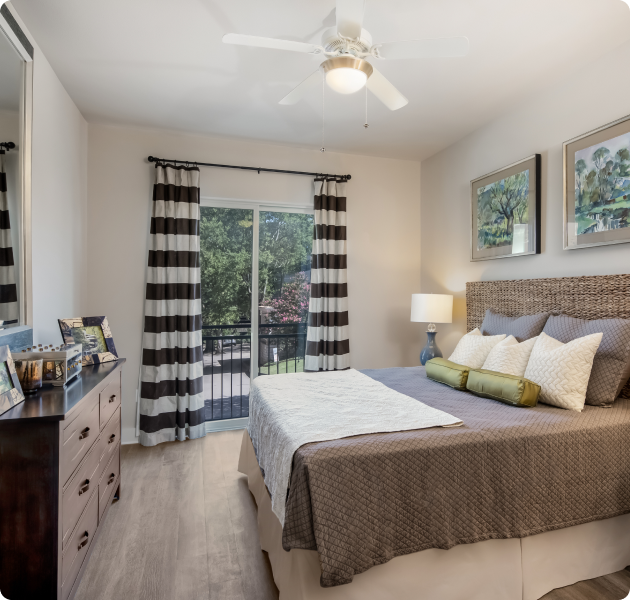 Apartment bedroom with ceiling fan and vanity mirror above a wood dresser next to sliding glass doors on wood floors at MAA Acklen luxury apartments in Nashville, TN