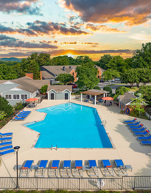 Aerial view of resort-style pool with a pergola, blue lounge chairs and orange sun umbrellas, table and chairs surrounded by lush trees at sunset at MAA Brentwood luxury apartments in Nashville, TN