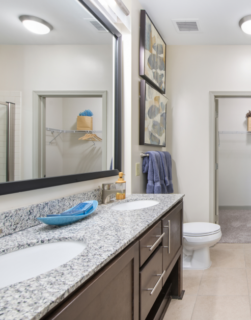 Modern designed bathroom with light fixtures above large framed vanity mirror, double sinks on granite countertops with dark brown cabinets next to a walk-in closet at MAA Charlotte Ave luxury apartments in Nashville, TN