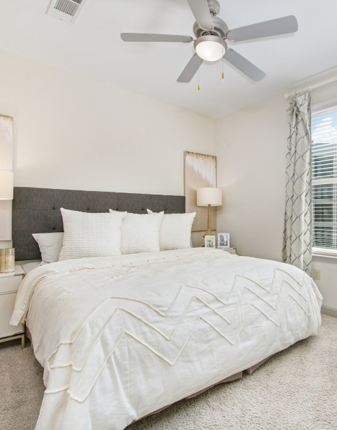 Carpeted bedroom with ceiling fan, night stands with lamps and white bedding with accent pillows next to a window with blinds and curtains at MAA Charlotte Ave luxury apartments in Nashville, TN