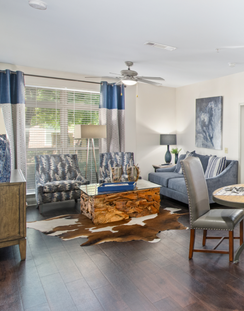 Spacious model living room with a ceiling fan, large picture window with blinds and curtains, a sitting area with a carved wood coffee table on wood floors at MAA Charlotte Ave luxury apartments in Nashville, TN