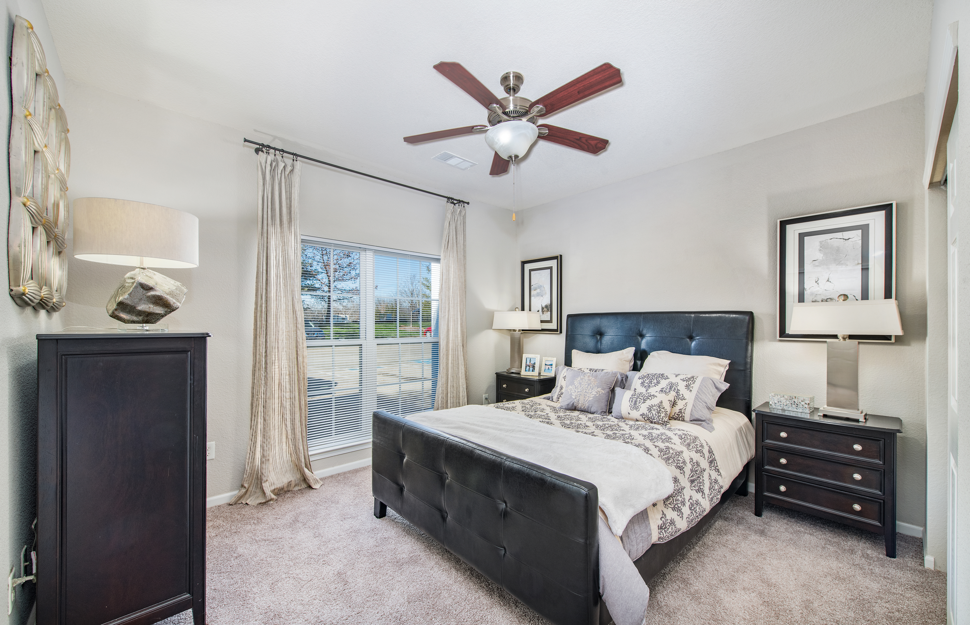 An apartment model bedroom with ceiling fan and wall paintings, tufted headboard queen bed with wood night stands next to a window covered with blinds and curtains at MAA Indian Lake luxury apartments in Hendersonville, TN