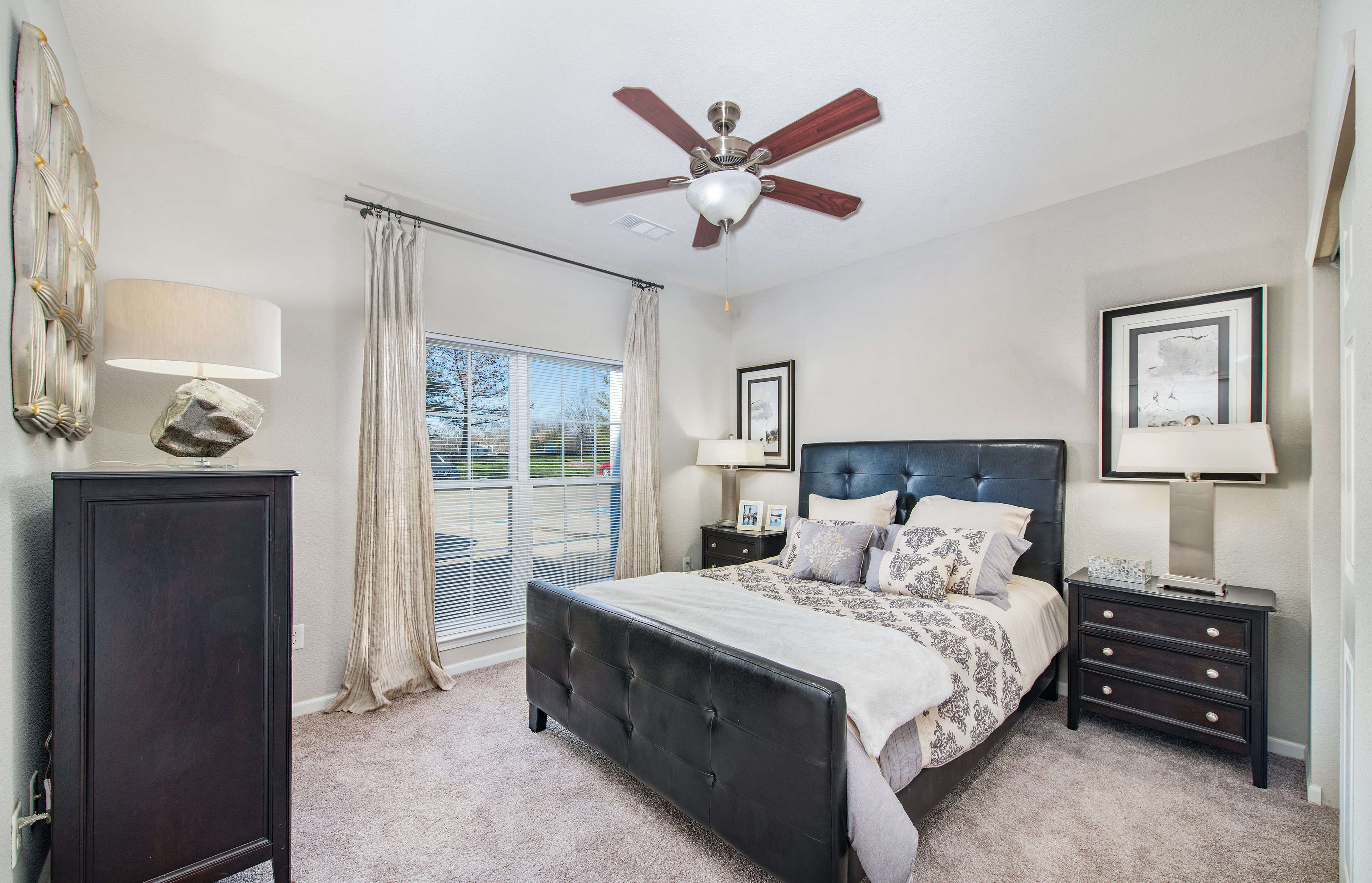 An apartment model bedroom with ceiling fan and wall paintings, tufted headboard queen bed with wood night stands next to a window covered with blinds and curtains at MAA Indian Lake luxury apartments in Hendersonville, TN