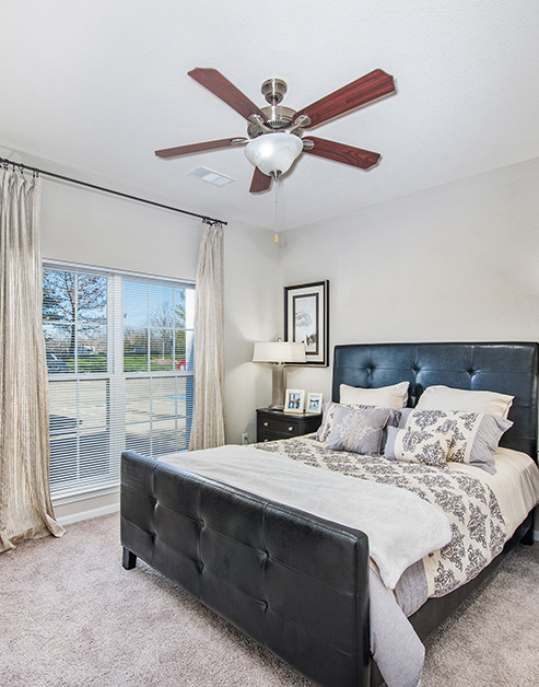 Carpeted bedroom with ceiling fan, bedside table with lamp next to the large window with blinds and curtains at MAA Indian Lake luxury apartments in Hendersonville, TN