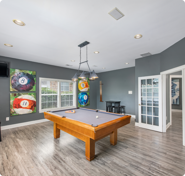 Spacious community clubhouse game room with wall art, hanging light fixtures over a pool table next to window with blinds and wood floors at MAA Monthaven Park luxury apartments in Hendersonville, TN