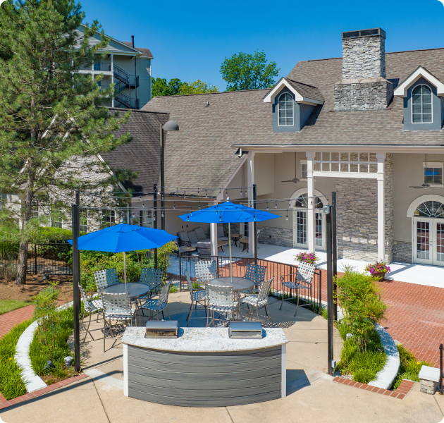 Aerial view of community outdoor grilling station area with patio tables, chairs and blue sun umbrellas next to clubhouse at MAA Nashville West luxury apartments in Nashville, TN