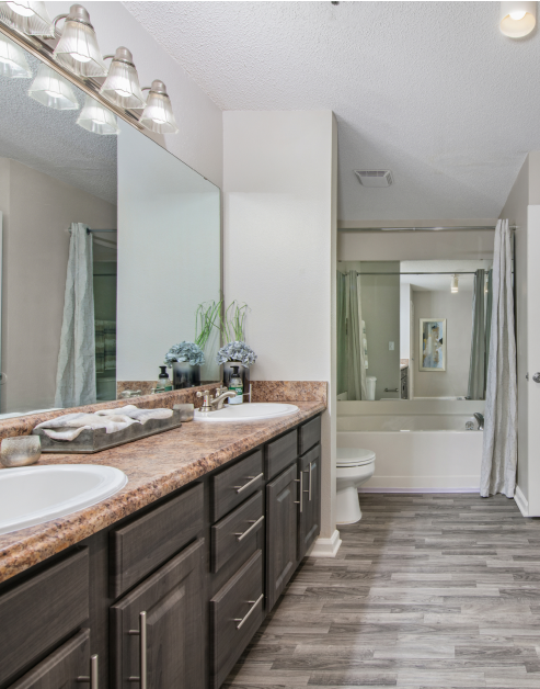 Modern bathroom with light fixtures above large vanity mirror and double sinks on brown countertops with dark grey cabinets and wood floors at MAA Nashville West luxury apartments in Nashville, TN