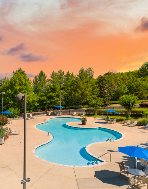 Sunset view of resort-style pool with lounge chairs and blue sun umbrellas with table and chairs surrounded by lush trees at MAA Nashville West luxury apartments in Nashville, TN