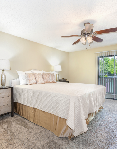 Bedroom with ceiling fan, nightstand with lamp and white bedding with accent pillows next to sliding glass door with vertical blinds at MAA Park luxury apartments in Hermitage, TN