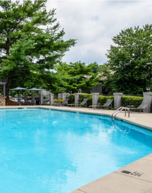 Gated swimming pool with sun umbrellas and lounge chairs surrounded by lush greenery at MAA Park luxury apartments in Hermitage, TN