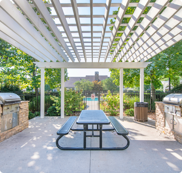 Community outdoor shaded pergola with grilling stations and a bench table, surrounded by lush trees and gated pool at MAA Sam Ridley luxury apartments in Smyrna, TN