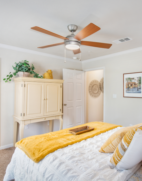 Bedroom with ceiling fan, wall painting and white bed with yellow blanket and accent pillows next to dresser cabinet at MAA Sam Ridley luxury apartments in Smyrna, TN