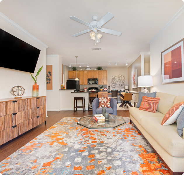 Apartment living room with a ceiling fan, wall-mounted TV above wooden cabinet and a beige couch next to a blue armchair and glass coffee table on colorful area rug on wood floors at MAA Onion Creek luxury apartments in Austin, TX