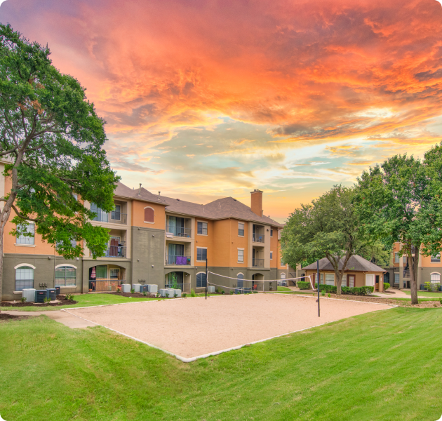 Community outdoor sand volleyball court surrounded grass and lush greenery with a sunset sky next to the apartment building at MAA Quarry Oaks luxury apartments in Austin, TX.