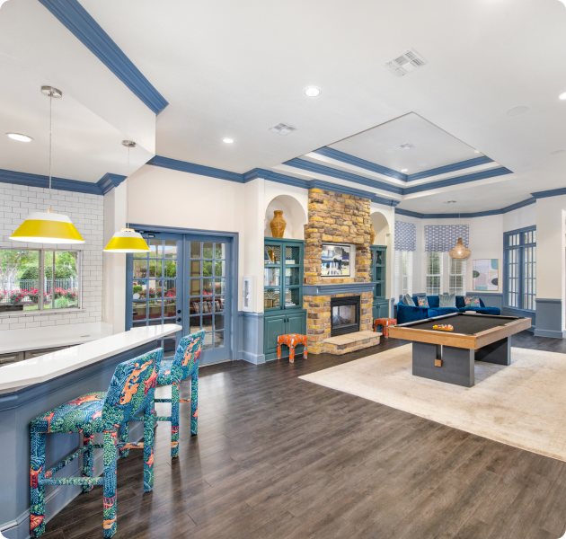 Spacious community clubhouse with blue-toned trim and beveled ceiling, hanging lights above a bar-top kitchen area with colorful bar chairs, pool table, and a brick fireplace next to double French door entrance at MAA Round Rock luxury apartments in Round Rock, TX.