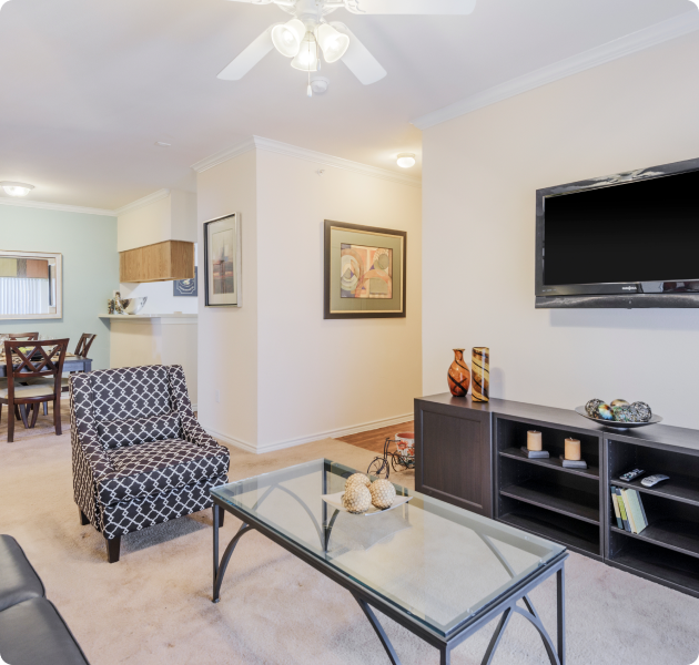 Apartment living room with ceiling fan, accent sofa chair with a coffee table next to a wall-mounted TV above cabinet next to dining area at MAA Sierra Vista luxury apartments in Round Rock, TX