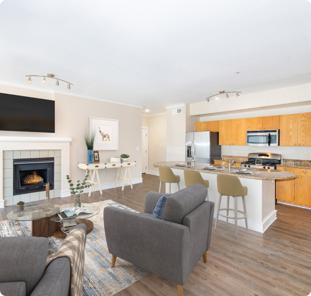 A model living room featuring an inviting sitting area with sofa chairs, hardwood floors, a kitchen, dining space, and a fireplace at MAA Abbey luxury apartments in Dallas, TX