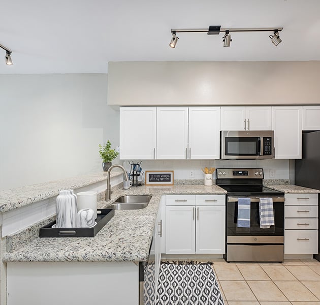 Modern apartment kitchen with track lighting, granite countertops and a double sink, white cabinets, stainless steel appliances and tile floors at MAA Addison Circle luxury apartments in Addison, TX