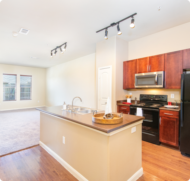 A luxurious apartment with an open kitchen with a center island and modern appliances, adjacent to living room with carpeted floors at MAA Bella Casita luxury apartments in Irving, TX