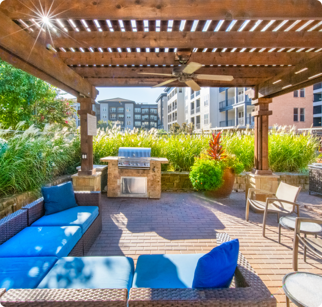 Outdoor seating area with a wicker-style patio sofa set and a barbecue grill station, surrounded by tall grasses and greenery at MAA Bella Casita luxury apartments in Irving, TX