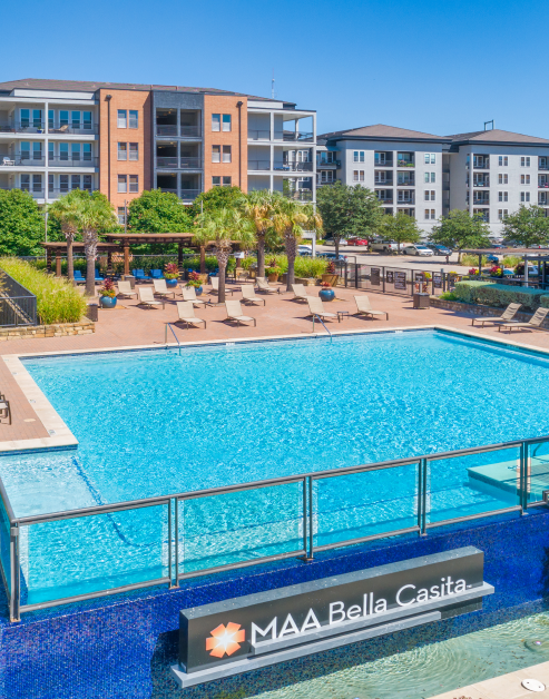 Stunning swimming pool with lounge chairs and sundeck surrounded by large trees and planters at MAA Bella Casita luxury apartments in Irving, TX