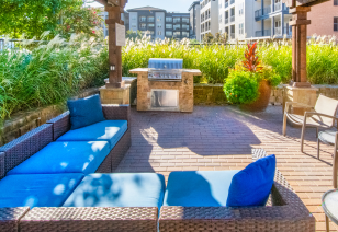 Poolside seating area with a wicker-style patio sofa set and a barbecue grill station, surrounded by tall grasses and greenery at MAA Bella Casita luxury apartments in Irving, TX