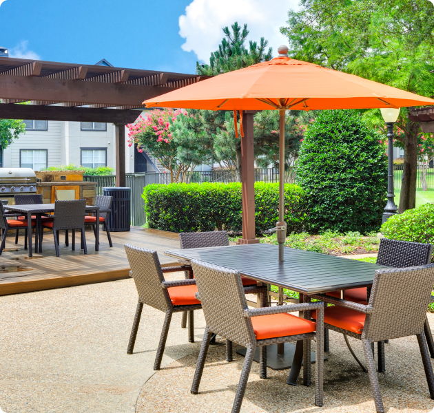 An outdoor community area with an orange sun umbrella and patio table with chairs next to a pergola on a wooden deck with a grill surrounded by trees at MAA Boulder Ridge luxury apartments in Roanoke, TX