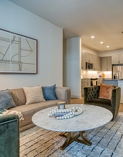 Luxury living area with a beige couch and a dark green velvet armchair next to a large round white marble coffee table on an area rug next to the kitchen at MAA Cathedral Arts luxury apartments in Dallas, TX